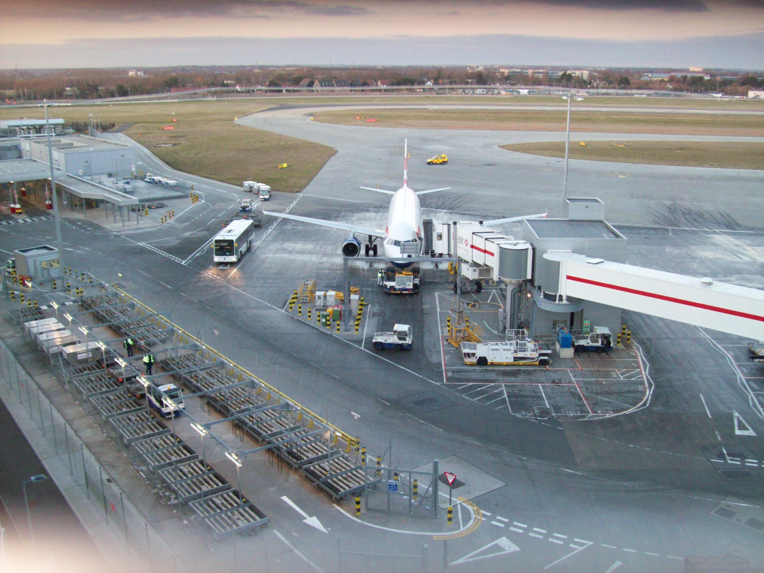 Avion de ligne à sa passerelle sur l’aire d’un aéroport, avec véhicules de piste, bus et marquages au sol, vu d’en haut.
