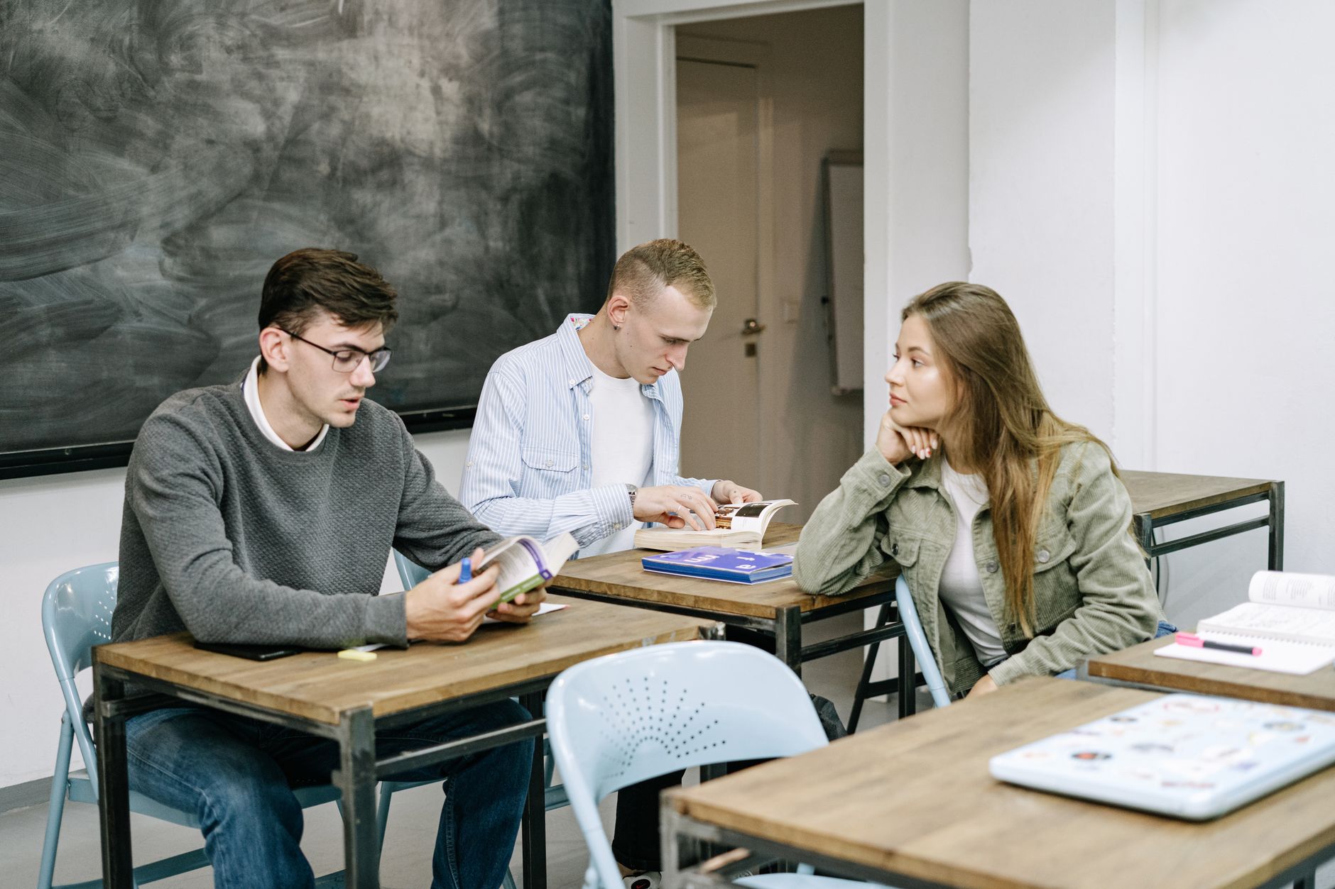 Trois jeunes adultes assis à des bureaux dans une salle de classe, deux lisent des livres, une femme les observe devant un tableau noir.